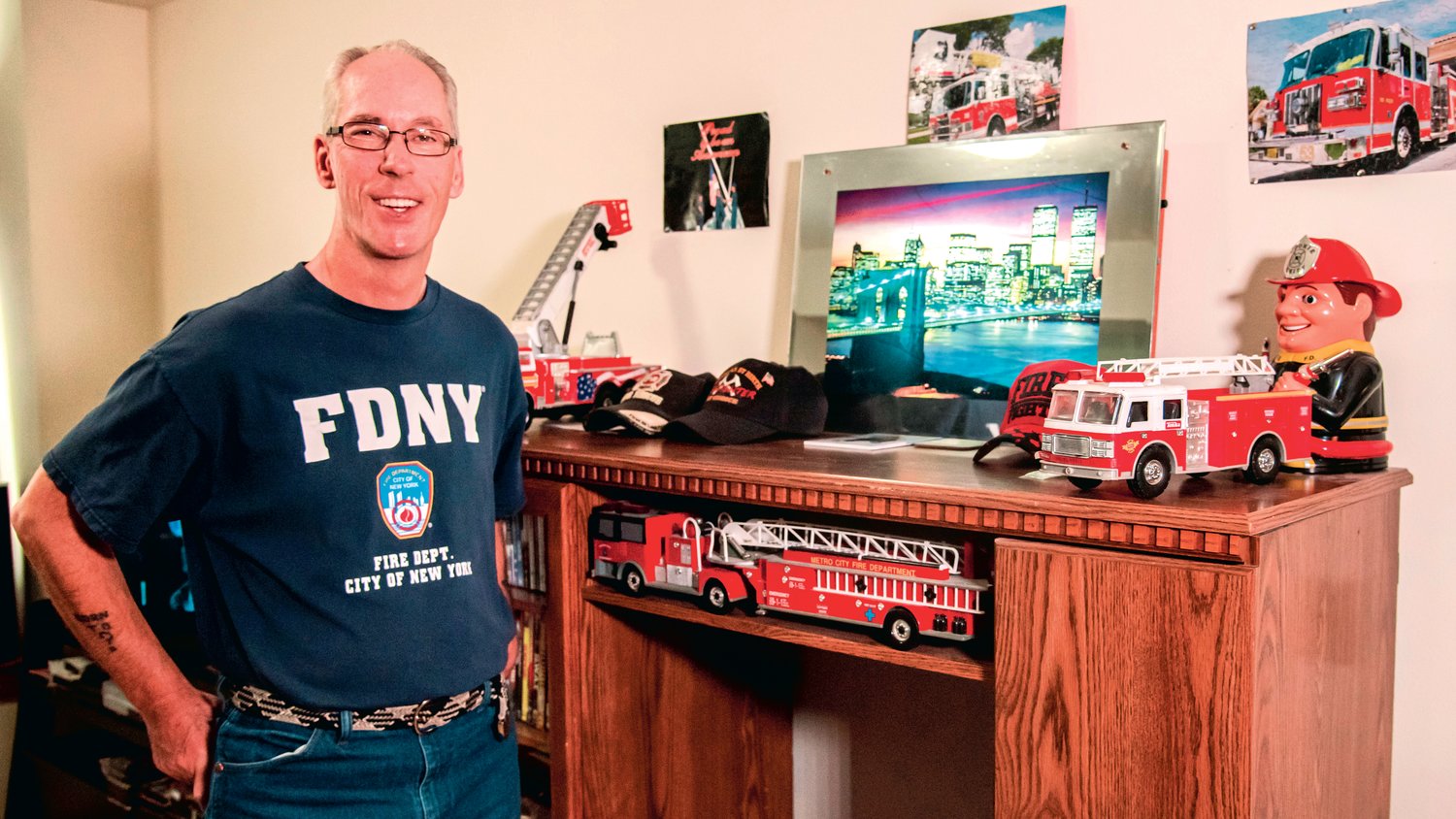 Chuck Tippett Jr. smiles and poses for a photo next to a collection of fire hats and engines displayed next to an illuminated photo of the New York City skyline in his Centralia residence. Tippett Jr. also sports his “FDNY” shirt he received while working as a firefighter in the city where he helped victims during the events that unfolded on September 11, 2001. "That's my boys right there," he said Saturday while describing the fire trucks displayed in his living room.